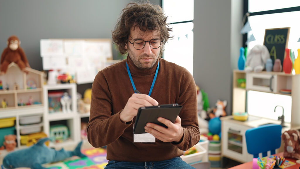 A male educator sitting in a kindergarten room, completing administration task on a tablet