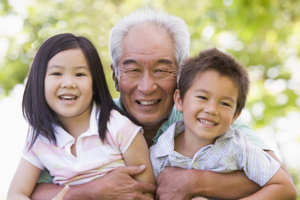 A grandfather smiling while holding two children. Grandparent Care is one of the categories for additional childcare subsidy ACCS