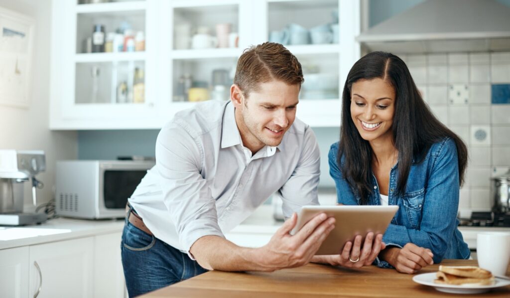 A smiling couple in a kitchen, looking at a tablet, perhaps enjoying an update shared by an early childhood teacher