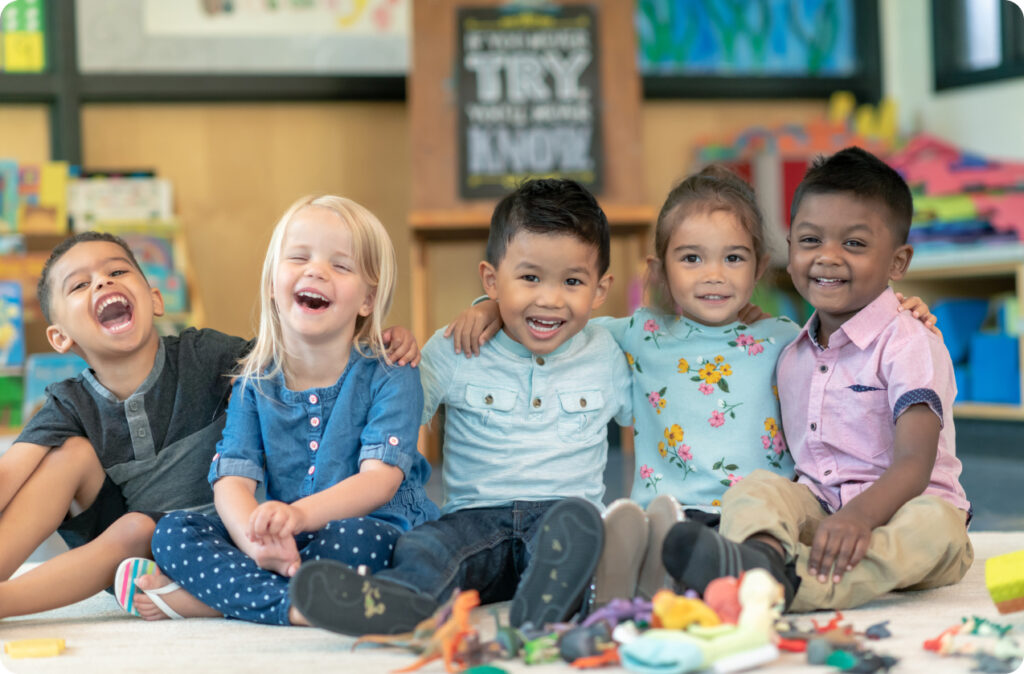 Five children of multiple ethnicities sitting and smiling with their arms around each other