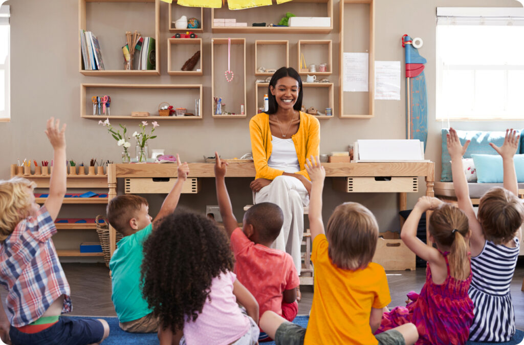 A smiling teacher looks at children sitting on the floor, with their hands raised ready to contribute, a sure sign of a great teacher in early learning