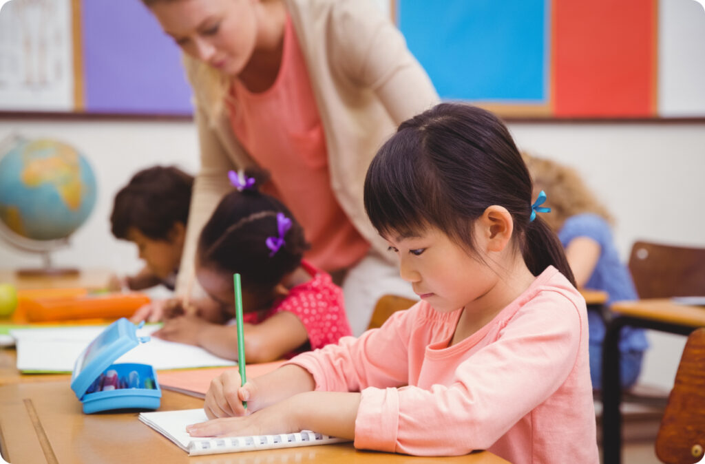 A young girl concentrates on writing as her teacher helps another student